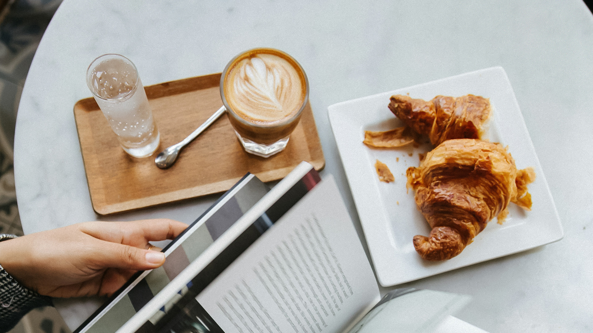 a person holding a tablet and a book next to a plate of croissants and a drink