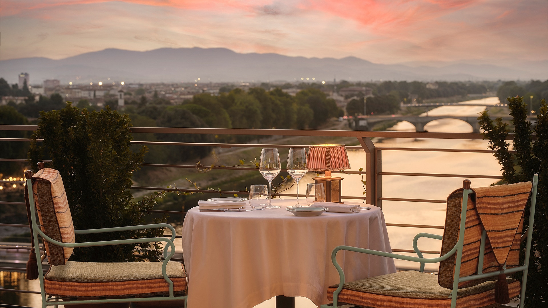 a table with wine glasses and a lamp on a balcony overlooking a river