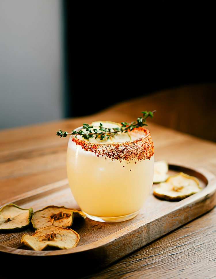 a glass of drink with a sprig of thyme on top of a wooden tray