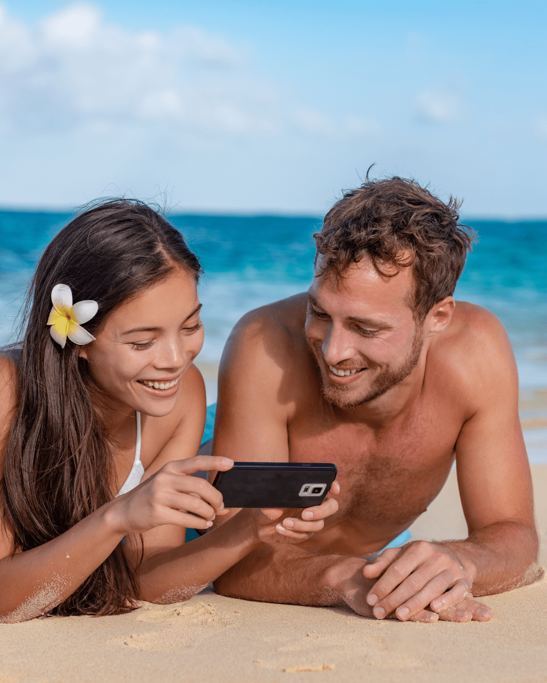 a man and woman lying on the beach looking at a cellphone