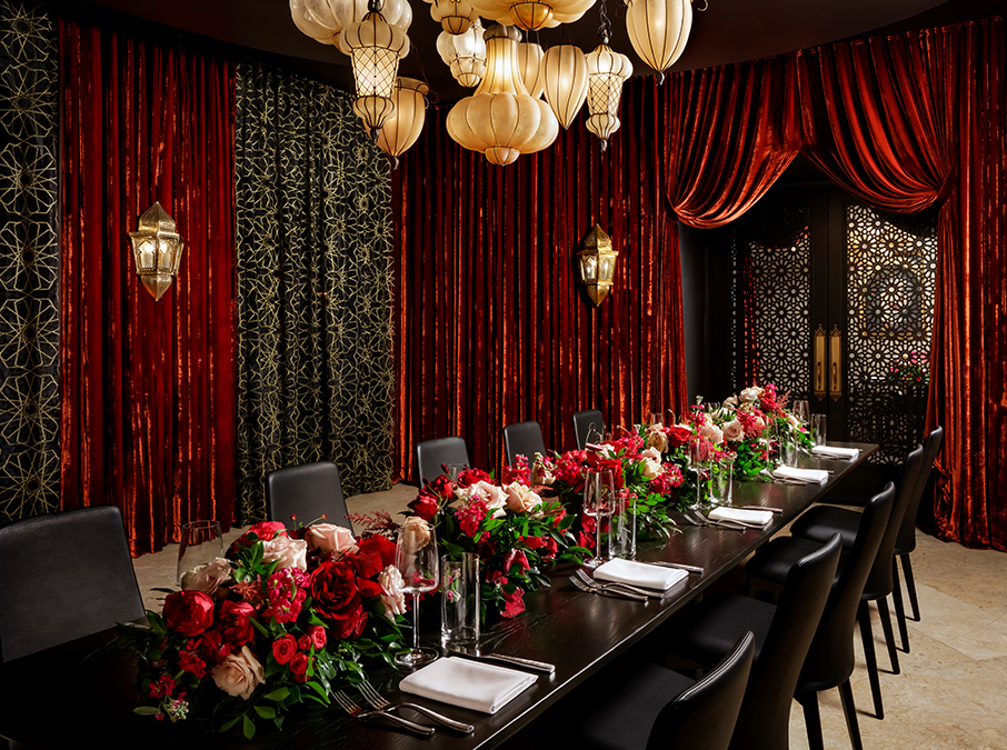 a long table with flowers and chairs in a room with red curtains