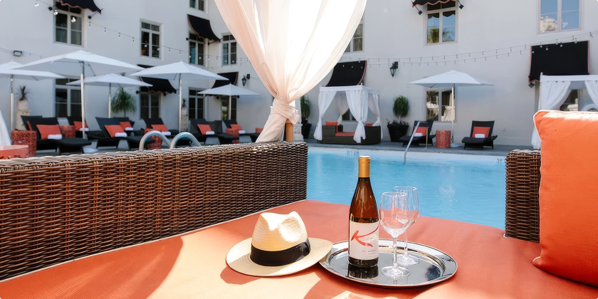 a wine bottle and glasses on a table with a hat and a pool in the background