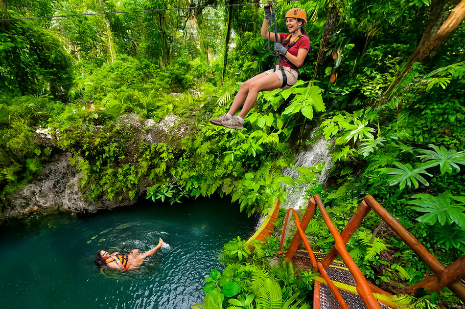 a woman in a helmet on a zip line above a man in a pool