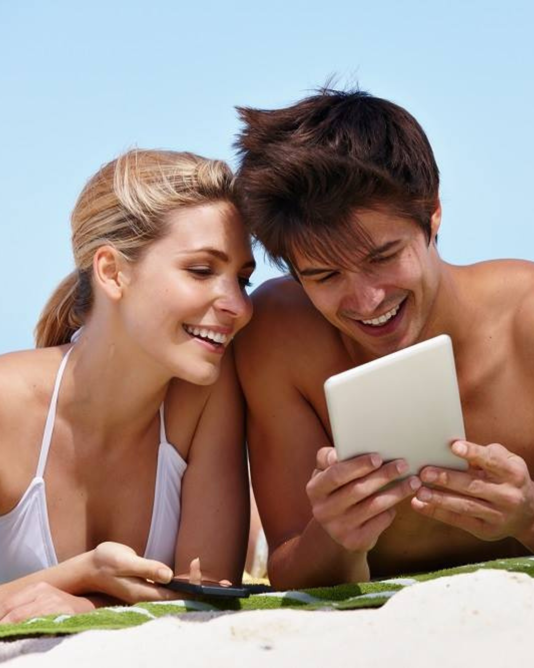 a man and woman lying on a towel looking at a tablet